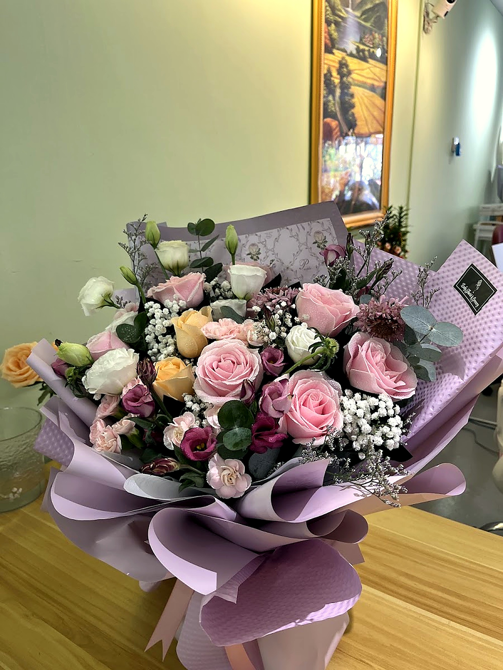 A floral bouquet of pink and peach roses, white lisianthus, and baby's breath wrapped in layered lilac paper