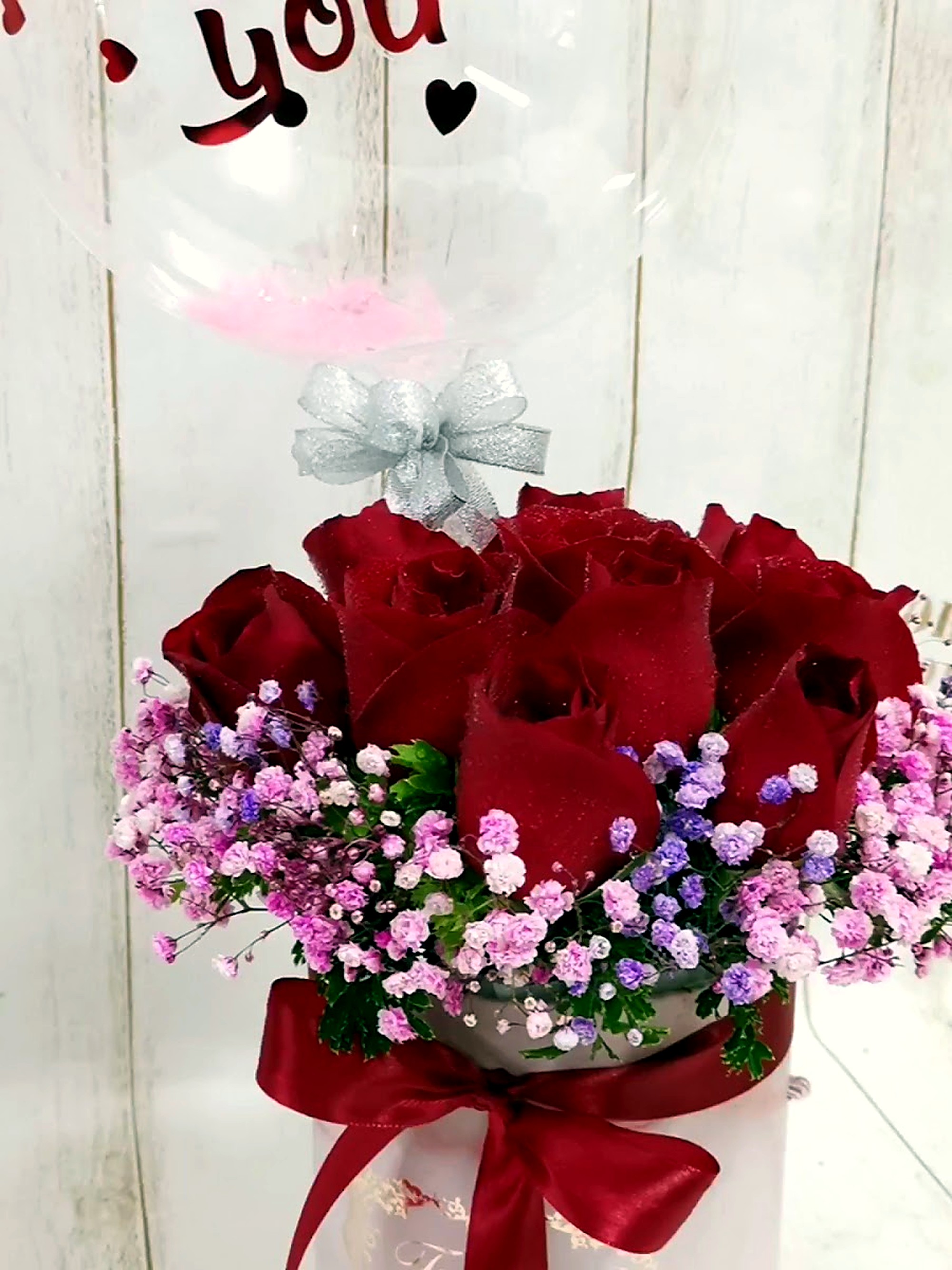 A floral arrangement of red roses and pink baby's breath in a gift box topped with a clear balloon